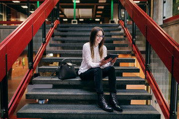 Smiling Caucasian female student with eyeglasses and brown hair sitting on the stairs in college...