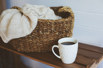 cozy winter interior details in white and brown tones. Basket with knitted sweater and cup of coffee on wooden shelf.