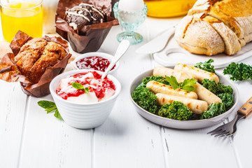 Breakfast served with muffins, grilled sausages, juice, fresh bread and parfait on white wooden table.