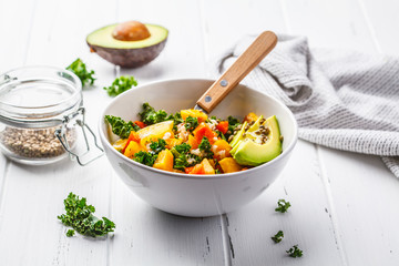 Vegan salad with rice, kale, baked pumpkin, carrots and avocado in white bowl on white wooden background.