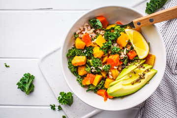Vegan salad with rice, kale, baked pumpkin, carrots and avocado in white bowl on a white wooden background.