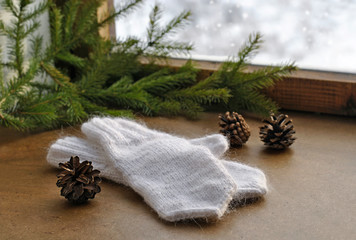Obraz na płótnie Canvas Warm homemade knitted white wool mittens, fir branches, cones and an old wooden winter window. Christmas or New Year card.