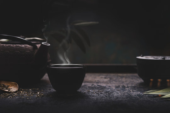 Dark tea background with black iron asian teapot and mug of hot tea with steam on table. Copy space for your design. Authentic vintage style. Traditional tea ceremony arrangement