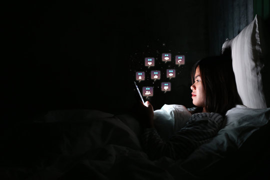 Asian Woman Using Smartphone For Checking Social Media With Icon Or Hologram At Night On The Bed In Dark Room, Using Smartphone In Dark May Be Causes Of Eye Pain Symptom