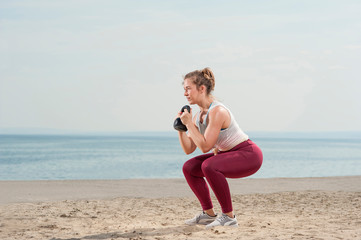 Young attractive woman dressed in pink leggings doing a squat with a kettle bell by the seaside. Fitness woman working out at the beach. Fitness Concept.