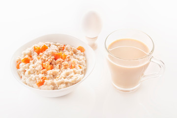 Oatmeal with pumpkin and nuts in a plate, a glass of tea, a boiled egg and a jug with milk on a white background. Close-up.