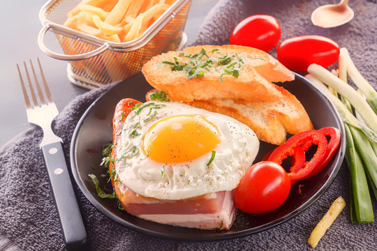 Fried egg with bacon in a black plate with fried pieces of bread, greens tomatoes and French fries on a gray wooden table. Close-up