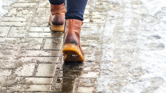 Walk On Wet Melted Ice Pavement. Back View On The Feet Of A Man Walking Along The Icy Pavement. Pair Of Shoe On Icy Road In Winter. Abstract Empty Blank Winter Weather Background