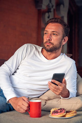 Man sitting on a terrace sofadrinking coffee and eating donuts while using cellphone.