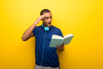 African american man with blue t-shirt on yellow background holding a book and surprised while...