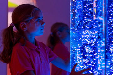 Child in therapy sensory stimulating room, snoezelen. Child interacting with colored lights bubble tube lamp during therapy session. - obrazy, fototapety, plakaty