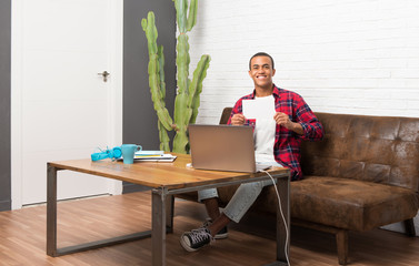 African american man with laptop in the living room holding an empty white placard for insert a...