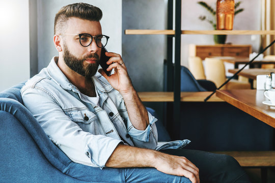 Portrait of young man in trendy glasses sitting in cafe and talking on cell phone. Young male entrepreneur has telephone conversations. Freelancer works remotely. Social communication.