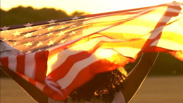 4K video clip of mixed race African American girl teenager female young woman holding an American USA Stars and Stripes flag in a wheat field at sunset or sunrise