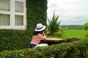 Young female sitting at the terrace of tree leaves covered country house, looking at the paddy field 