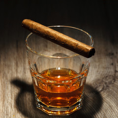 A glass of alcoholic beverage with ice cubes and a cigar on a wooden table. Whiskey in glass