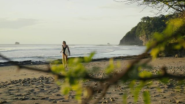 SLOW MOTION: Close up shot of a young beautiful woman on a sandy beach running with a surfboard in her hands. From blurry to clear shot.
