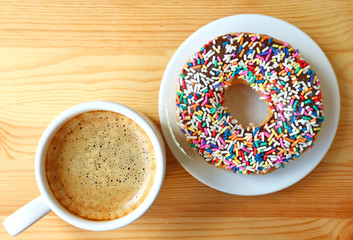 Top view of a cup of hot coffee and a donut topped with colorful sprinkles served on wooden table 