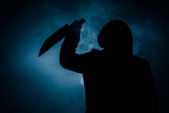 A dangerous hooded man standing in the dark and holding a knife. Face can not be seen. Committing a crime concept. Selective focus