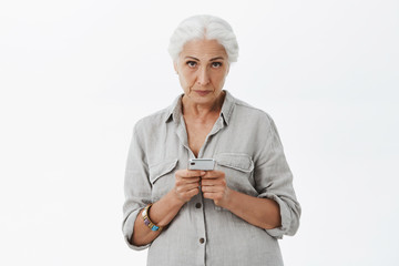 Indoor shot of calm and serious-looking irritated senior woman with gray hair looking with contempt form under forehead feeling annoyed kid interfering her while using smartphone over white wall