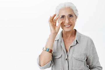 Waist-up shot of charming kind and nice granny in sight glasses touching rim of eyewear and smiling joyfully helping grandson prepare for exams posing joyful and delighted over grey wall