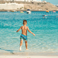 Active European boy in a striped swimming shorts is running towards the sea. He is having fun spending his holidays on the seashore.