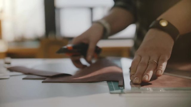 Beautiful close-up shot of skilled female artisan hands cutting big leather piece on table with knife and triangle ruler