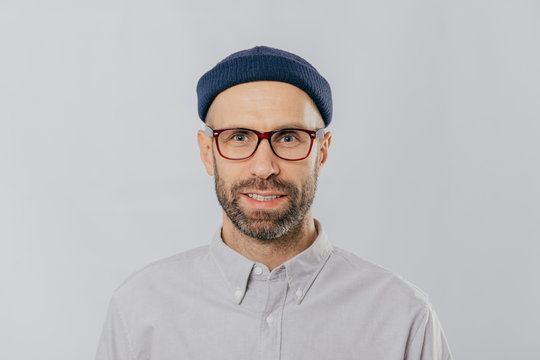 Headshot of handsome unshaven man wears transparent glasses, headgear, formal shirt, looks directly at camera, isolated over white background. Caucasian male with stubble dressed in fashionable outfit