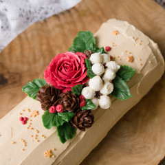 Christmas sponge roll decoreted with cream flowers - poinsettia, cotton, rose, cones - on a wooden dish on wooden background. Chocolate yule log cake. xmas or new year theme