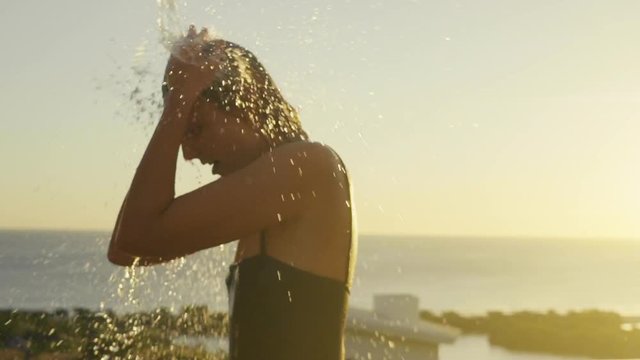 A young blonde woman in black swimsuit is enjoying a tropical outdoor shower in sunset. She is happy.
