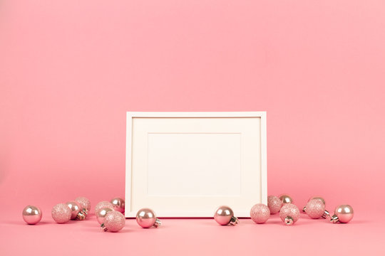 Mock up with horisontal white picture frame on trendy pastel light pink background with christmas ornaments. Greeting card and pink Christmas baubles