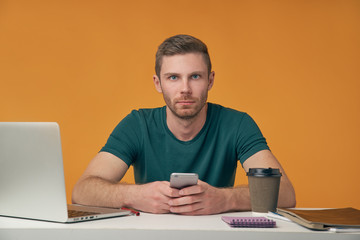 a young man with a beard in a t-shirt sitting at a Desk with a laptop and a Cup of coffee holding a smartphone and look carefully at the camera
