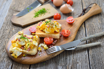 Deftige Rühreier mit Schinkenwürfel und Schnittlauch auf Toastbrot heiß serviert – Hearty scrambled eggs with bacon and chives served hot on toasts
