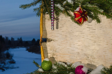 Новогодняя композиция в виде поздравительной открытки из березовой коры украшенной еловыми ветками и новогодними игрушками на фоне зимнего пейзажа.