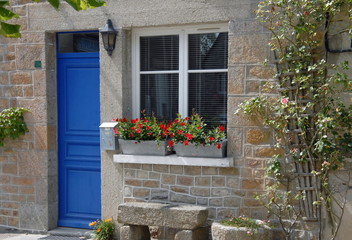 Fototapeta na wymiar Ville de Genêts, maison en pierre, porte bleue et fleurs au bord de la fenêtre, banc en pierre, département de la Manche, France