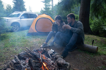 Obraz premium couple sitting near camp fire and drinking tea. tent and suv on background