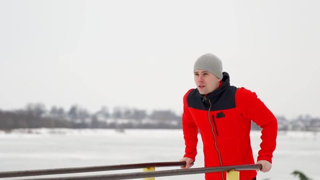 A young handsome man in a sports jacket and black trousers in winter on an open platform performs otshimaniya on parallel bars. Slow motion. Winter landscape