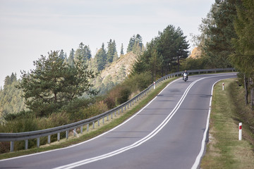 Fototapeta premium Alone motorcyclist in black leather outfit riding cruiser motorbike along twisted road on background foggy distant green woody hills under morning sky.