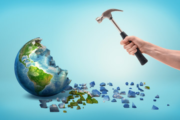A male hand holds a metal hammer near a semi-broken Earth globe with small pieces fallen out of it.