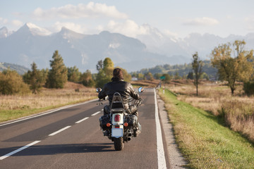 Fototapeta na wymiar Back view of biker in black leather jacket riding motorcycle along road on blurred background of beautiful mountain range with snowy peaks, moving vehicles on bright sunny summer day.