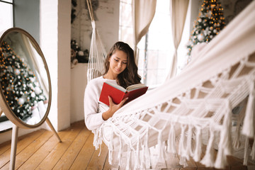 Beautiful dark-haired girl dressed in pants and sweater reads a book lying in a hammock next to the...