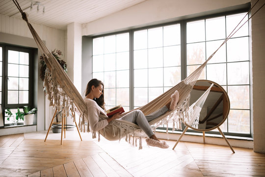 Dark-haired girl dressed in pants, sweater and warm slippers reads a book lying in a hammock in a cozy room with wooden floor and panoramic windows and a round mirror on the floor