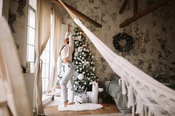 Charming girl dressed in white sweater and pants stands next to the New Year tree in front of the window and stretches up  in a cozy decorated room  with New Year's wreath