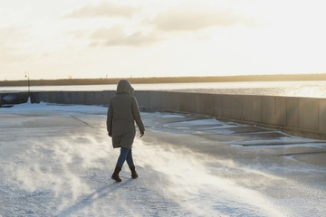 Back view woman in a winter jacket with a hood going on the embankment of a freezing river in...