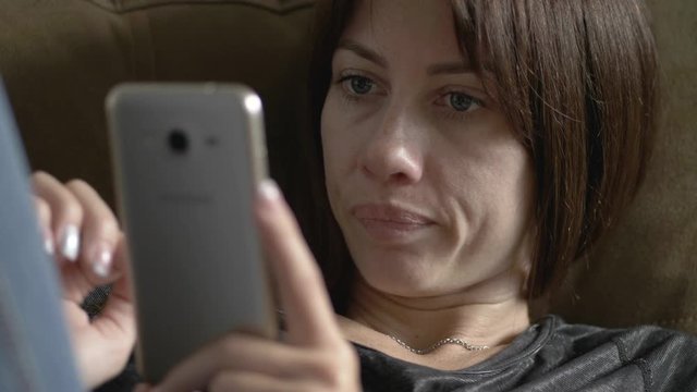 Close up shot of adult woman using smartphone at sofa in home, scrolling on touch screen, emotionally reacting, tapping, choosing options, ponders, making decisions.