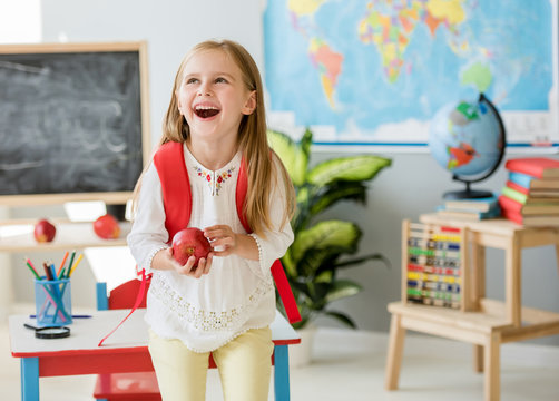 Little laughing blond girl holding an apple in the school classroom