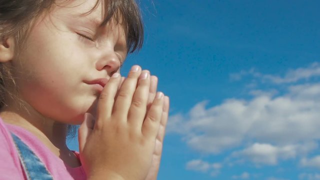 Portrait of a praying child. Cute little girl prays against the background of the sky with clouds. A child in prayer.