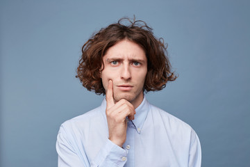 Thoughts and doubts concept. Man with long messy hair thoughtful face, holding fingers on cheek and chin, gazing camera. Guy with untidy hairstyle has doubts, copy space, blue background.