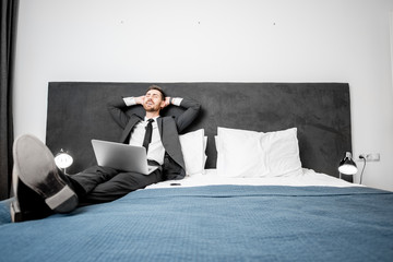 Relaxed businessman lying on the bed during his work with laptop at the hotel room during a business trip