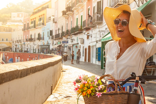 Young woman with bike at Ponza Island harbor pier in Italy. Tourist with large hat, fashion shirt and colorful skirt. Basket with wine and flowers in front of shops and boats.